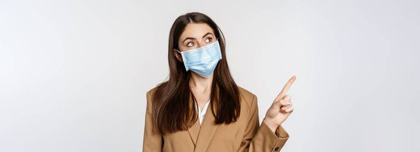 Coronavirus and people concept. Portrait of business woman at workplace wearing face mask, pointing finger left at logo, company banner, white background. - Φωτογραφία, εικόνα
