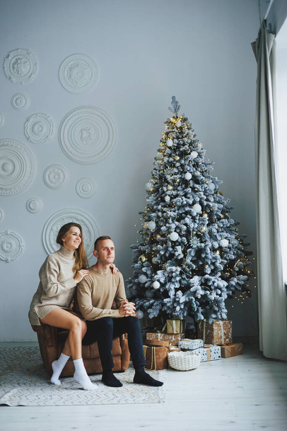 A couple in love on New Year's Eve in a cozy home environment near the Christmas tree. New Year's love story. Beautiful couple in sweaters posing in studio. A guy and a girl celebrate the new year - Photo, Image