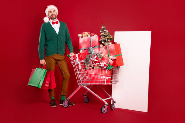 Full body photo of gentleman guy wear green cardigan red bowtie near supermarket cart stack gifts near white banner isolated on red color background. - Fotoğraf, Görsel