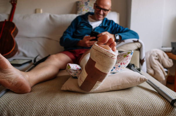 Hombre lesionado con el pie vendado usando un teléfono inteligente. - Foto, Imagen