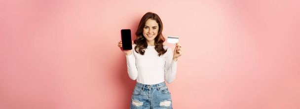 Online αγορές και καταστήματα έννοια. Νεαρή όμορφη γυναίκα δείχνει οθόνη του κινητού τηλεφώνου και πιστωτική κάρτα, συνιστώντας την εφαρμογή πληρωμής, στέκεται σε ροζ φόντο. - Φωτογραφία, εικόνα