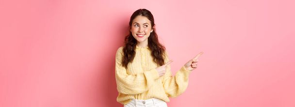 Young happy woman smiling, pointing and looking aside at left copyspace, showing advertisement, standing against pink spring background. - Photo, Image