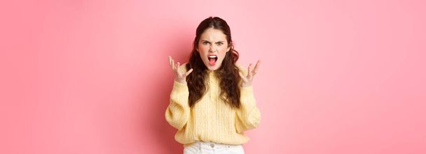Angry and pissed-off girl screaming, shaking hands and grimacing furious, stare with outraged annoyed face at camera, standing against pink background. - Photo, Image