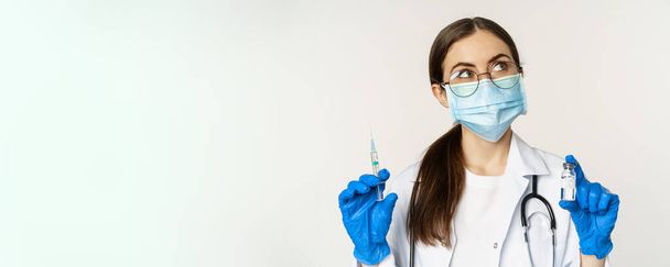 Covid-19 vaccination et concept de soins de santé. Jeune médecin en masque médical montrant la seringue et le vaccin contre le coronavirus variante omicron, debout sur fond blanc. - Photo, image