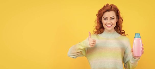 Γυναίκα απομονωμένη προσωπογραφία, πανό με χώρο αντιγραφής. ευτυχισμένη κοκκινομάλλα γυναίκα με σγουρά μαλλιά κρατώντας σαμπουάν ή conditioner εμφάνιση αντίχειρα, κομμώτρια - Φωτογραφία, εικόνα