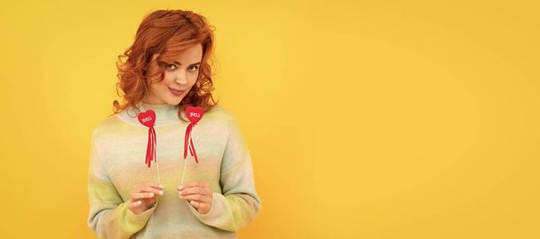 Woman isolated face portrait, banner with copy space. cheerful flirty redhead girl with red heart sticks on yellow background, love - Photo, image