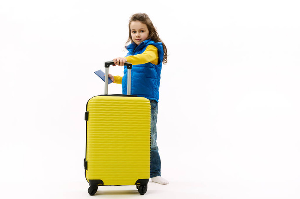 Full size traveler little child girl, in blue jacket, denim, holds a yellow suitcase and boarding pass, isolated on white background. Tourist travel abroad rest getaway Air flight trip journey concept - Fotó, kép