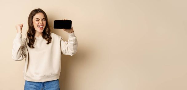 Happy girl showing mobile phone horizontal screen and celebrating, chanting with joyful face, standing over beige background. - 写真・画像