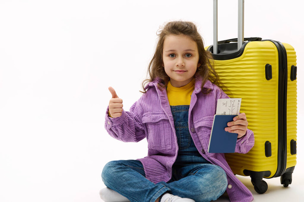 Szczęśliwa dziewczynka w fioletowej koszuli i dżinsowych kombinezonach, siedząca w pobliżu żółtej walizki, trzymająca kartę pokładową i pokazująca kciuk przed kamerą, wainująca podczas lotu, odizolowana na biało. Dzieci wyjeżdżają za granicę - Zdjęcie, obraz