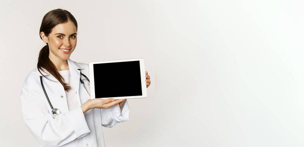 Kép a nő orvos, női egészségügyi dolgozó bemutató online orvosi honlap, digitális tabletta képernyő és mosolygós, álló fehér kabátban fehér háttér. - Fotó, kép