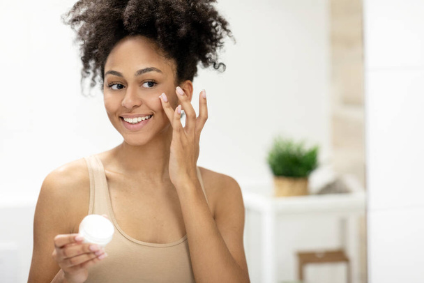 Mosolygó afro-amerikai nő arcápoló krémet használ, miközben fogja az üveget és tükörbe néz. Kiváló minőségű fénykép - Fotó, kép