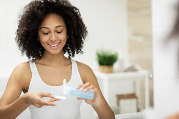 A mulher negra limpa a pele facial com água micelar e algodão, enquanto desfruta da rotina de cuidados com a pele no banheiro moderno em casa. Foto de alta qualidade - Foto, Imagem