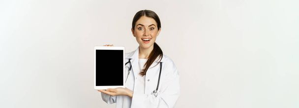 Imagen de la mujer doctora, trabajadora de la salud mostrando el sitio web médico en línea, la pantalla digital de la tableta y sonriendo, de pie en bata blanca sobre fondo blanco. - Foto, Imagen
