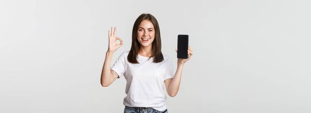 Fille brune souriante agréable montrant l'écran du smartphone et montrant un geste correct dans l'approbation. - Photo, image