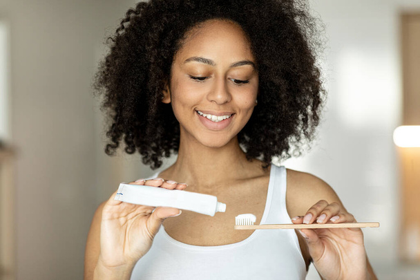 A dark-skinned woman smiles and squeezes toothpaste on an ecological wooden brush. High quality photo - Foto, afbeelding