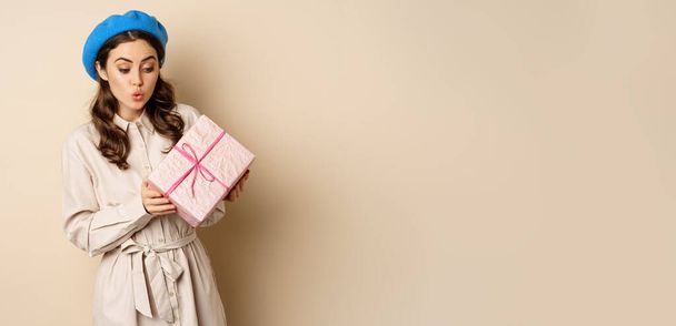 Holidays and gifts concept. Beautiful girl receive gift box and looking happy, holding pink wrapped present with joyful face expression, beige background. - Photo, Image