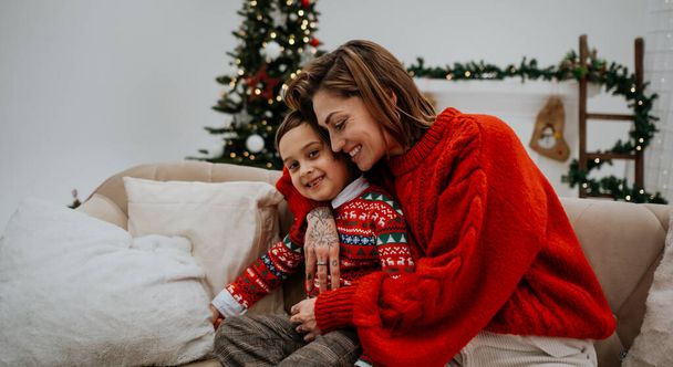 Joven madre con el pelo oscuro abrazándose con su hijo cerca del árbol de Navidad en la habitación decorada para las vacaciones. Concepto de ambiente familiar y navideño - Foto, Imagen