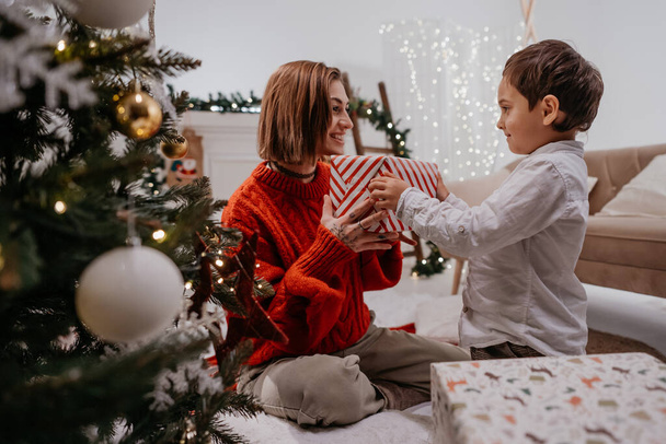 Un ragazzo adorabile che fa un regalo a sua madre sotto l'albero di Natale. Single genitore femminile trascorrere del tempo con il figlio a casa e festeggiare il nuovo anno insieme - Foto, immagini