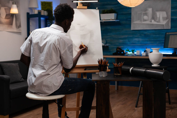 Afrikkalainen amerikkalainen henkilö taiteellisia kykyjä laittaa viimeistely koskettaa lyijykynä piirustus maljakko. Lahjakas nuori mies luo potin ääriviivat kankaalle maalausteline kotona käsitöitä atelier. - Valokuva, kuva