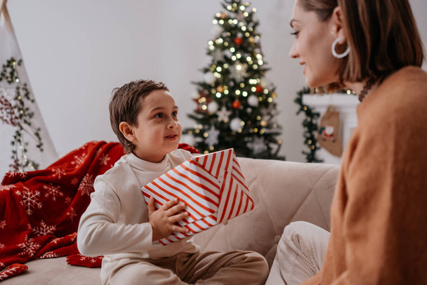 Carrying woman giving Christmas present to the boy. Single mother spending time with son at home and celebrating New Year together - Φωτογραφία, εικόνα