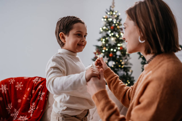 Femme brune regardant son fils avec un sourire tendre tout en célébrant le Nouvel An à la maison. Mignon garçon se réjouissant avec sa mère célibataire - Photo, image