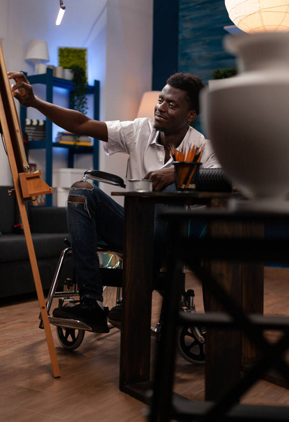 Kunstenaar met een handicap maakt vaas op doek. Happy Afro-Amerikaanse kunst student het creëren van kunstwerk met gekleurde potloden in huis studio. Rolstoelgebruiker die nieuwe artistieke vaardigheden leert. - Foto, afbeelding
