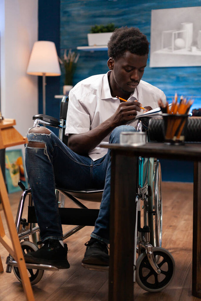 Mężczyzna rysunek profesjonalny użytkownik wózka inwalidzkiego pisanie notatek z papieru i ołówka na biurku w sypialni. Afrykański amerykański artysta żyjący w stanie zdrowia co ręcznie rysowane szkic z kolorowym ołówkiem. - Zdjęcie, obraz