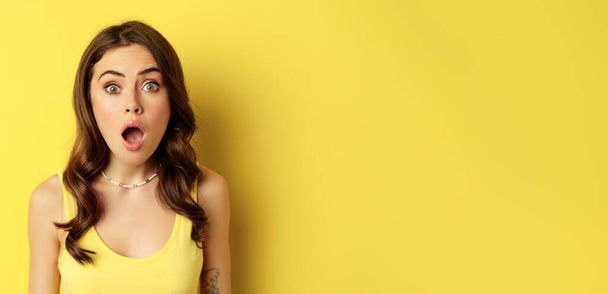 Close up portrait of stylish brunette girl looking amazed, wow face, showing excitement and surprise, impressed by something, standing over yellow background. - Photo, Image