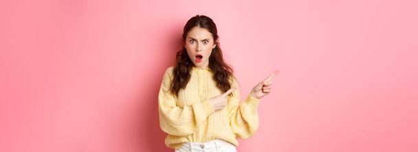 Shocked and frustrated young brunette woman, gasping and staring startled at camera, pointing fingers left at copy space, standing against pink background. - Foto, Imagen