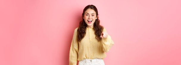Young brunette woman laughing, smiling amazed and pointing at camera, choosing you, standing against pink background. Copy space - Foto, afbeelding