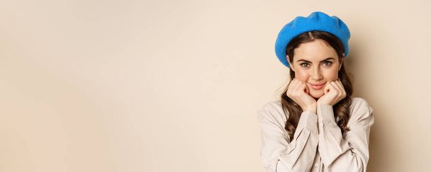 Primer plano retrato de mujer joven femenina en sombrero francés de moda, posando lindo y romántico, haciendo tonta cara coqueta y sonrisa tierna, fondo beige. - Foto, Imagen