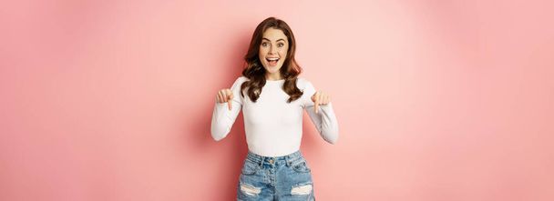 Enthusiastic beautiful and stylish girl showing advertisement, pointing fingers down at announcement and smiling, pink background. - Fotografie, Obrázek