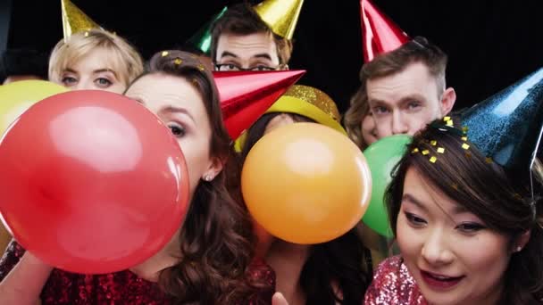 Party, confetti and people with balloons in studio for festive celebration, social event and birthday on black background. Friends, fun and portrait of men and women blowing balloon with party hats. - Footage, Video