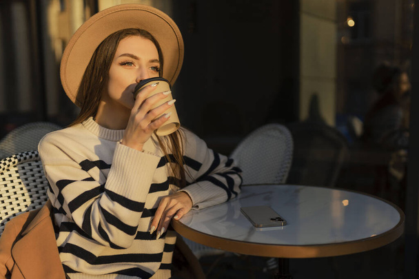 krásná usmívající se žena ve stylovém oblečení sedí u stolu v klobouku a svetru, romantická šťastná nálada, čeká na přítele na rande v kavárně, jaro-letní módní trend, pití kávy. - Fotografie, Obrázek