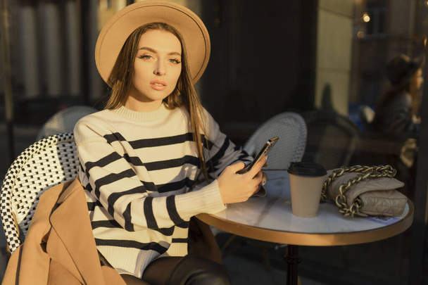 Attractive cheerful brunette girl in trench coat with coffee to go happily using cellphone outdoor.  - Photo, Image