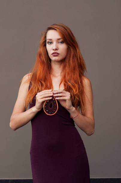 Portrait of beautiful slim redhead girl in wine boho chic dress and handmade white dreamcatcher feather talisman pendant - Фото, зображення