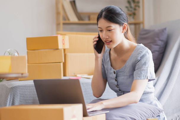 Mosolygó gyönyörű ázsiai nő KKV vállalkozó Vállalkozástulajdonos gazdaság kartondoboz termékek és ellenőrizze az ügyfelek megrendeléseit a laptopok otthon, koncepció KKV szállítás online. - Fotó, kép