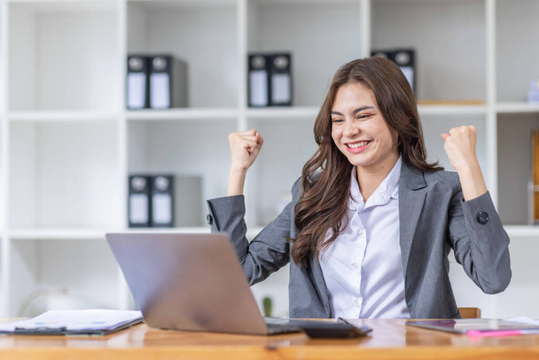 Szczęśliwy podekscytowany sukces azjatycki bizneswoman triumfuje ze smartfonem i laptopem w miejscu pracy. - Zdjęcie, obraz
