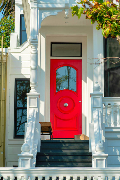 Červené malované přední dveře se skleněným oknem a šedými schody s tradičním bílým zábradlím a malebnou přední verandou s visící rostlinou ve skvrnitém poledním slunci. Předměstské odpoledne. - Fotografie, Obrázek