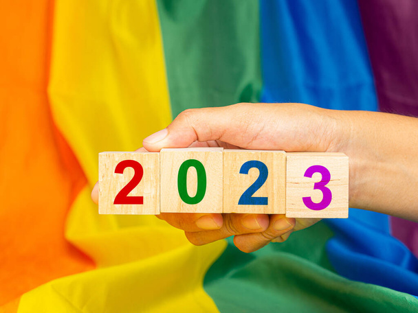 Dřevěné kostky s písmeny 2023 v ruce s duhovou vlajkou nebo pozadím vlajky LGBT. Fotografie zblízka. Koncept genderové rozmanitosti - Fotografie, Obrázek