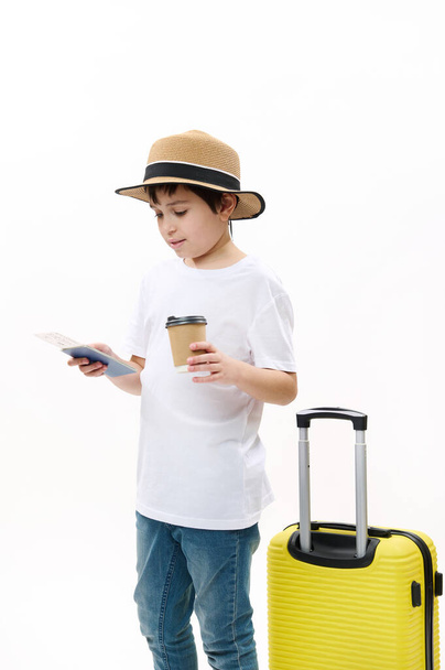 Uroczy nastolatek w niebieskich dżinsach, luźnej koszulce i słomkowym kapeluszu z żółtą walizką, trzyma gorący napój na wynos w jednorazowym papierowym kubku i patrzy na bilet lotniczy, odizolowany na białym tle - Zdjęcie, obraz