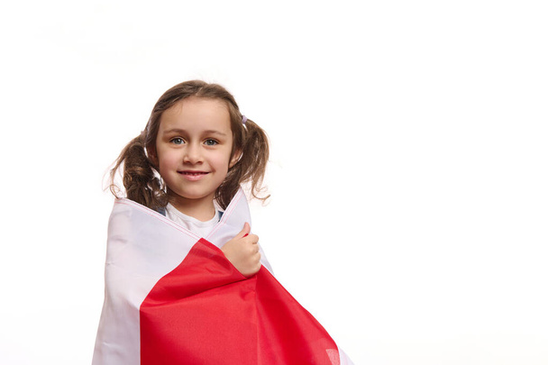 Petite fille européenne avec deux jolies queues de cheval enveloppant le drapeau de Pologne, sourire joyeux sourire aux dents en regardant la caméra, isolé sur fond blanc. Célébration de la fête de l'indépendance polonaise, 11 novembre - Photo, image