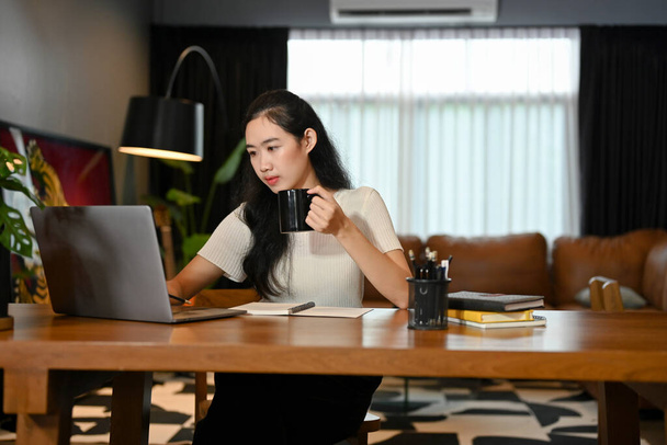 Szakmai fiatal ázsiai nő szabadúszó vagy üzletasszony kortyolgatja a kávét, és koncentrál a feladatára laptop számítógép. - Fotó, kép