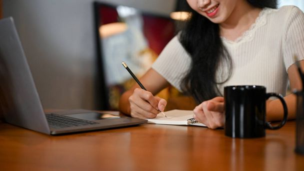 Jolie jeune étudiante asiatique qui fait ses devoirs dans son salon, écrit quelque chose sur son livre scolaire et utilise un ordinateur portable. image recadrée - Photo, image