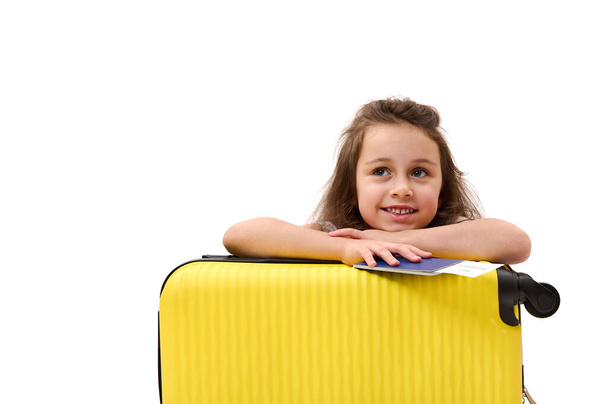 Schattig klein passagiermeisje, met instapkaart en koffer, een weekendje naar het buitenland, glimlachende tandenglimlach, dromerig wegkijkend naar advertentieruimte. Toerisme Tourney Luchtvaart concept - Foto, afbeelding