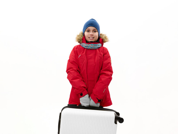 Středovýchodní teenager v červené bundě s kožešinovou kapucí, s modrým kufrem, izolovaný nad bílým pozadím s volným reklamním prostorem. Odpočinek Útěk Journey Cestování a Air letový koncept - Fotografie, Obrázek