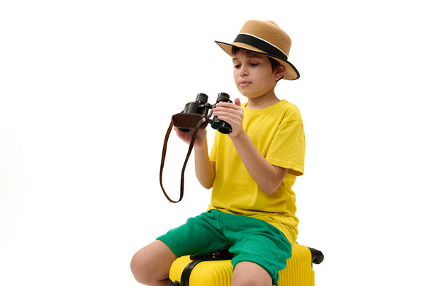 Entzückender frühpubertärer Junge - junger Touristenreisender in gelbem T-Shirt, grüner Sommerhose und Strohhut, sitzend auf einem Koffer mit Fernglas in der Hand, isoliert über weißem Hintergrund. Freier Anzeigenplatz - Foto, Bild