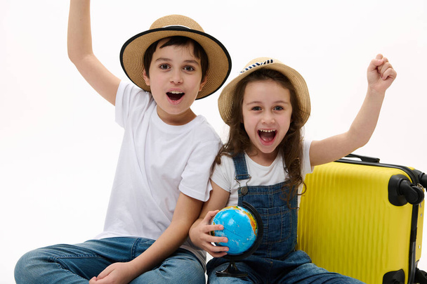 Niños alegres apretando los puños de la felicidad, disfrutando de los próximos viajes al extranjero, sentados contra una maleta amarilla con globo mapa de la Tierra, sobre fondo blanco y sonriendo alegremente mirando a la cámara - Foto, Imagen