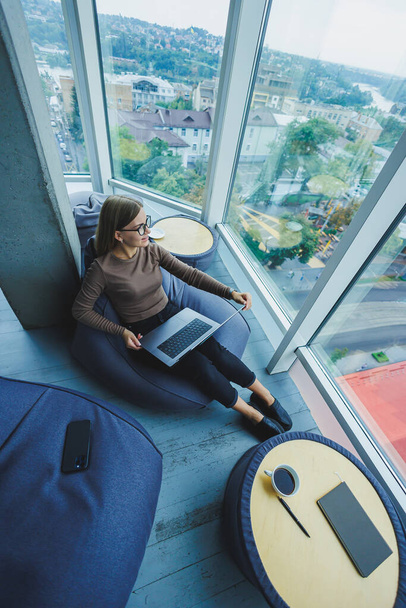 Красивая студентка работает на ноутбуке, сидя на кресле-бобовом мешке в современном творческом кабинете на фоне панорамных окон. Концепция прерывания времени - Фото, изображение