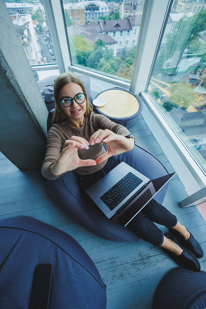 Una bella studentessa lavora su un computer portatile mentre siede su una sedia a sacco in un moderno ufficio creativo sullo sfondo di finestre panoramiche. Break time concept - Foto, immagini